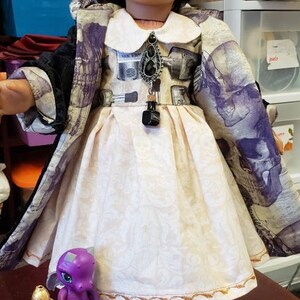 May include: A doll wearing a cream-colored dress with a floral pattern and a black and purple skull-print coat. The doll has dark hair and is wearing earrings. Accessories include a purple toy dragon and glass jars.