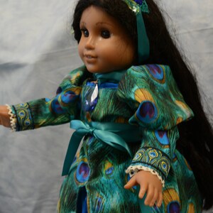 May include: A doll wearing a teal blue dress with a peacock feather pattern. The dress has a teal blue sash and the doll is wearing a green and blue peacock feather headband.