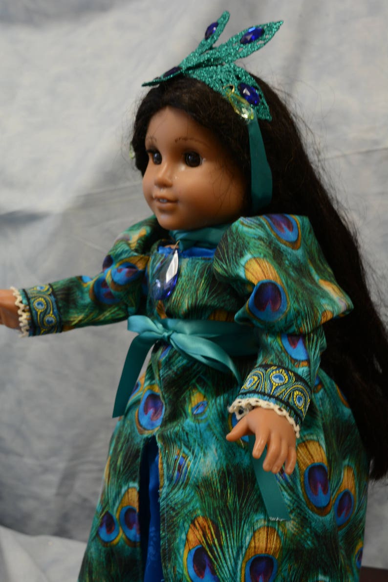 May include: A doll wearing a teal blue dress with a peacock feather pattern. The dress has a teal blue sash and the doll is wearing a green and blue peacock feather headband.