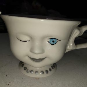 May include: A white ceramic teacup with a face design. One eye is winking, the other is open with a blue iris. The cup has a handle and a decorative base with small, silver-colored dots. The face has a gray smile.