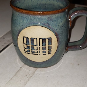 May include: A blue and brown ceramic mug with a white circle on the side. The circle has the word "DUM" written in black letters inside.
