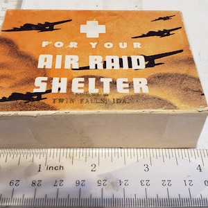 May include: A cardboard box with a white cross and the text "FOR YOUR AIR RAID SHELTER" printed on the top. The box is decorated with a brown and orange background and black silhouettes of airplanes.