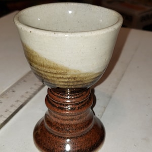 May include: A small, white ceramic cup with brown and black glaze sits on a brown and black ceramic pedestal. The cup has a rounded shape and a wide rim.