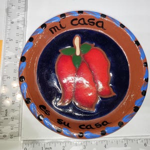 May include: Hand-painted ceramic plate with a red chili pepper design. The plate has a brown rim, a blue and brown border, and the words "mi casa es su casa" in black script. The plate measures approximately 6 inches in diameter.