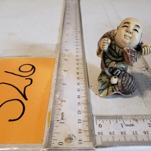 May include: A small, off-white figurine of a smiling man carrying a turtle on his back and holding a small package. The figurine is on a white surface with a ruler and an orange tag with the number "326" written on it.
