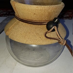 May include: A glass coffee maker with a cork and leather outer layer. The coffee maker has a brown leather strap with a black toggle.