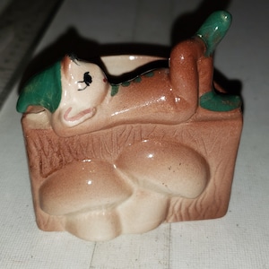 May include: A ceramic figurine of a green-hatted elf lying on a brown mushroom-shaped planter. The planter has a brown and white design.