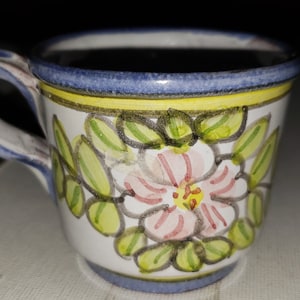 May include: A small white ceramic mug with a blue rim and a yellow band. The mug is decorated with a pink flower and green leaves.