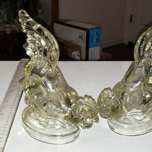 May include: Two clear glass angel figurines with wings outstretched. The angels are sitting on a base and are facing each other.