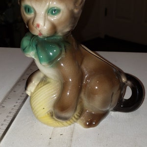 May include: A brown and white ceramic cat figurine with green eyes and a green bow. The cat is holding a yellow yarn ball.