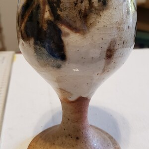May include: A ceramic goblet with a white glaze and brown and black abstract designs. The goblet has a wide base and a tall, slender stem.