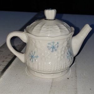 May include: A white ceramic teapot with a lid, decorated with blue snowflake designs. The teapot is approximately 3 inches tall.