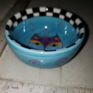 May include: A small, hand-painted ceramic bowl with a light blue interior and exterior. The bowl features a cat face design in purple, yellow, and green. The rim is decorated with a black and white checkered pattern.