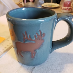May include: A teal ceramic mug with a brown elk silhouette. The mug has a rounded handle and a slightly textured surface. The interior is a darker teal color.