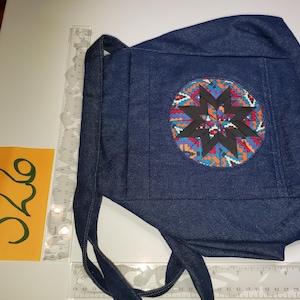 May include: A dark blue denim bag with a decorative circular patch featuring a starburst design in black, red, blue, and orange. The bag has a handle and a tie closure. A ruler and a card with the number "526" are also visible.