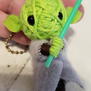 May include: A handmade Yoda-inspired keychain. The figure is crafted from green yarn, with black eyes and small green ears. It wears a gray felt cloak and holds a green lightsaber. A gold-colored chain is attached.