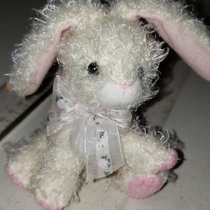 May include: A white plush bunny with pink ears and a white bow with green floral print. The bunny is about 3 inches tall.