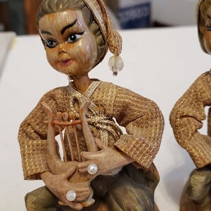 May include: A wooden figurine of a person playing a lyre. The figurine is wearing a gold and brown robe and a hat with a pearl. The figurine is sitting on a brown base.