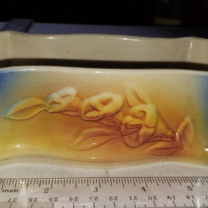 May include: A white ceramic planter with a blue and yellow glaze. The planter has a floral design with a branch of yellow flowers and green leaves.