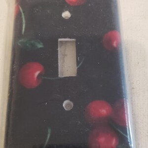 May include: A black light switch cover with a cherry print. The cherries are red and have green stems and leaves.