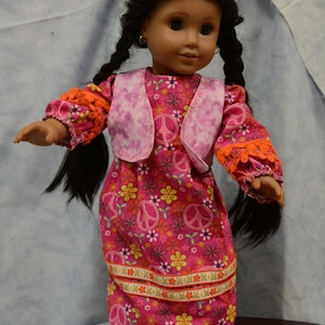 May include: A doll with long black braids wearing a pink and orange dress with peace signs and flowers. The doll is wearing pink shoes.