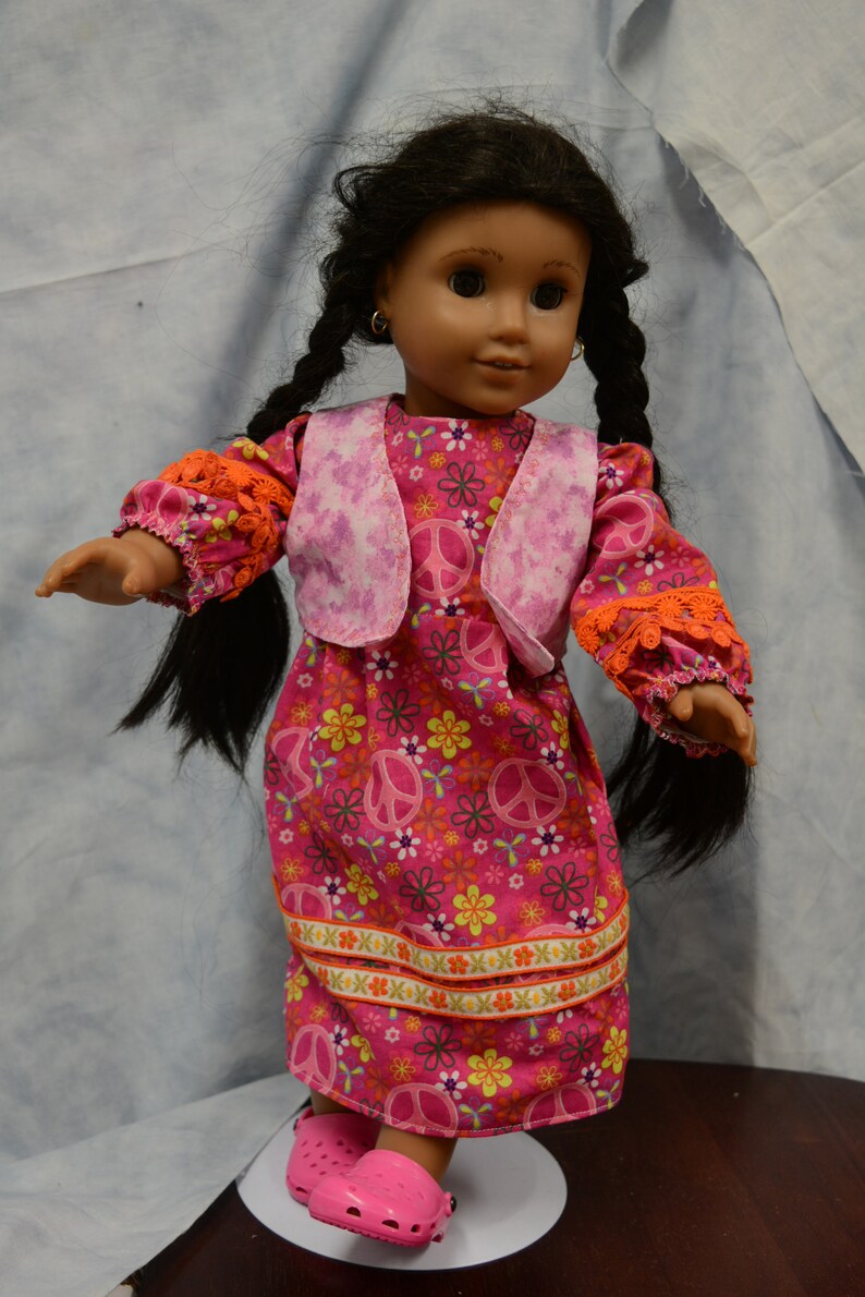 May include: A doll with long black braids wearing a pink and orange dress with peace signs and flowers. The doll is wearing pink shoes.