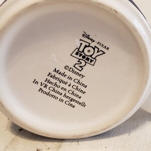 May include: White ceramic mug with a black and white image of the Toy Story 2 logo. The text on the mug reads "Disney Pixar Toy Story 2" and "&copy;Disney". The mug also has the text "Made in China" in black text.