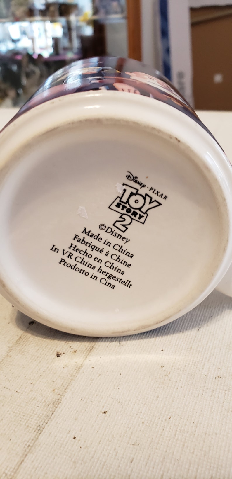 May include: White ceramic mug with a black and white image of the Toy Story 2 logo. The text on the mug reads "Disney Pixar Toy Story 2" and "&copy;Disney". The mug also has the text "Made in China" in black text.