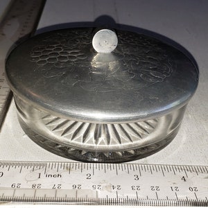 May include: A silver round trinket box with a decorative grape design on the lid and a fluted base. The box is approximately 2 inches in diameter.