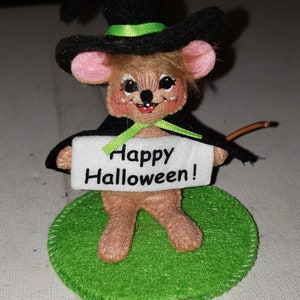 K&ouml;nnte beinhalten: Eine Halloween-Dekoration mit einer Maus, die als Hexe verkleidet ist. Die Maus tr&auml;gt einen schwarzen Hut mit gr&uuml;nem Band und einen schwarzen Umhang. Sie h&auml;lt ein Schild mit der Aufschrift "Happy Halloween!" und steht auf einem gr&uuml;nen Filzkreis.