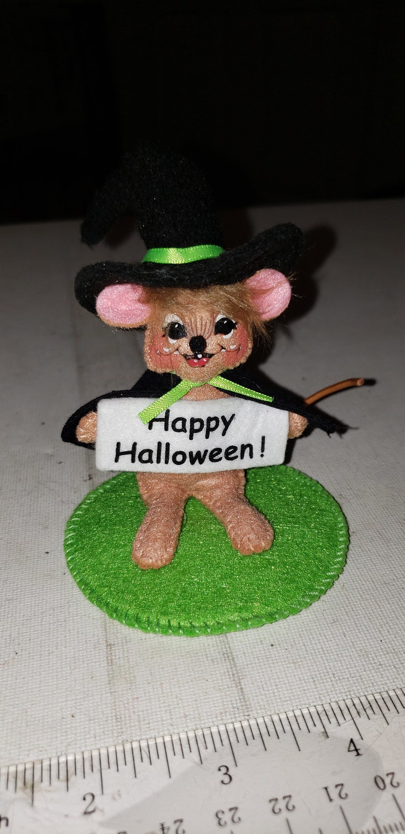 K&ouml;nnte beinhalten: Eine Halloween-Dekoration mit einer Maus, die als Hexe verkleidet ist. Die Maus tr&auml;gt einen schwarzen Hut mit gr&uuml;nem Band und einen schwarzen Umhang. Sie h&auml;lt ein Schild mit der Aufschrift "Happy Halloween!" und steht auf einem gr&uuml;nen Filzkreis.