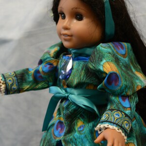 May include: A doll wearing a teal blue dress with a peacock feather pattern. The dress has a teal blue sash tied at the waist. The doll is wearing a teal blue headband with a sparkly butterfly design and blue jewels.