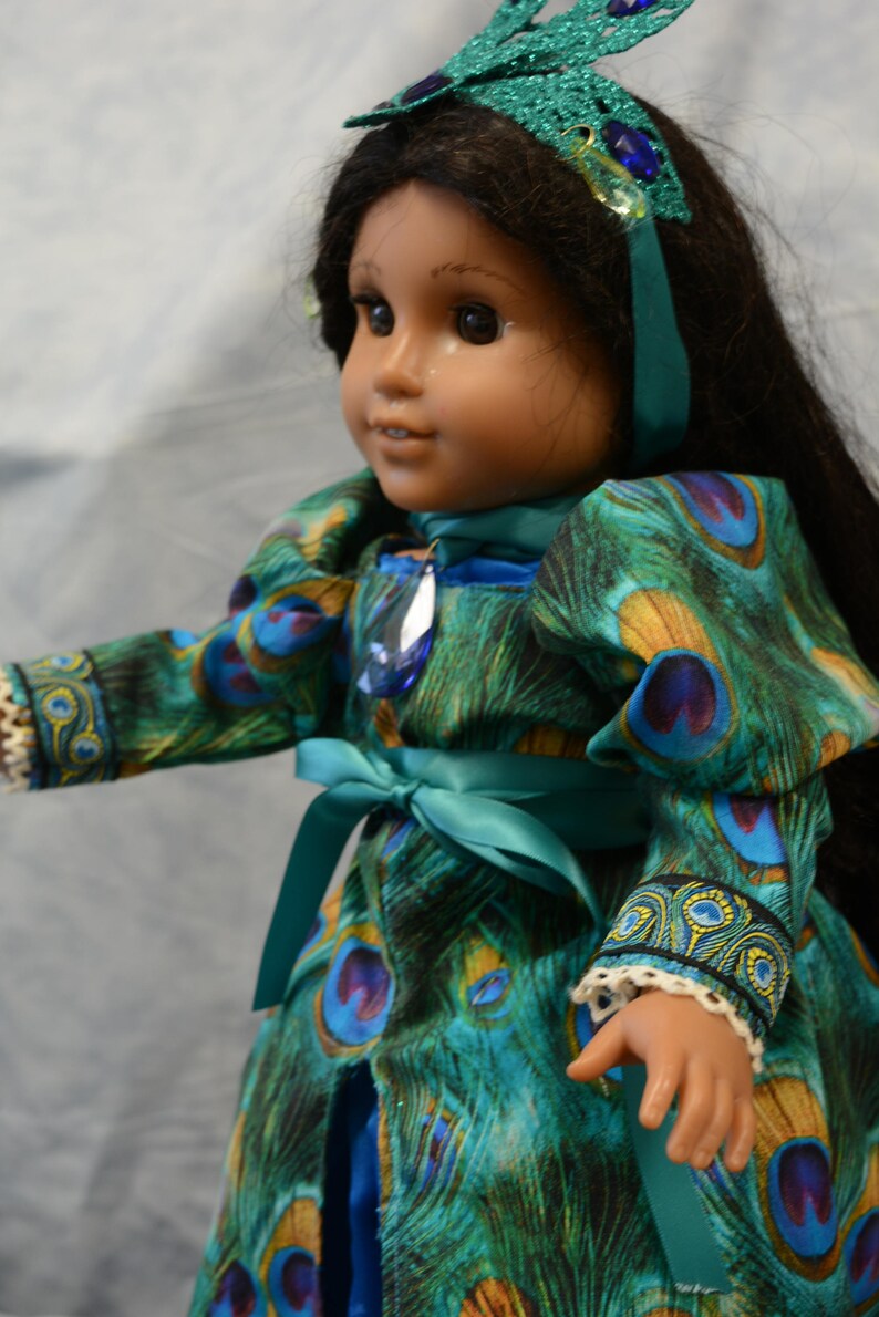 May include: A doll wearing a teal blue dress with a peacock feather pattern. The dress has a teal blue sash tied at the waist. The doll is wearing a teal blue headband with a sparkly butterfly design and blue jewels.