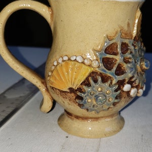 May include: A beige ceramic mug with a white rim and a handle. The mug is decorated with blue and brown gears, yellow and white seashells, and white dots.