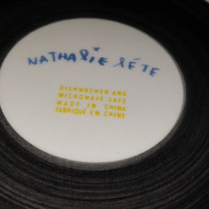 May include: A white ceramic plate with black concentric circles around the edge. The center of the plate has the text "NATHALIE RETE" in blue and yellow text. The text "Dishwasher and Microwave Safe Made in China Fabrique en Chine" is printed in yellow.