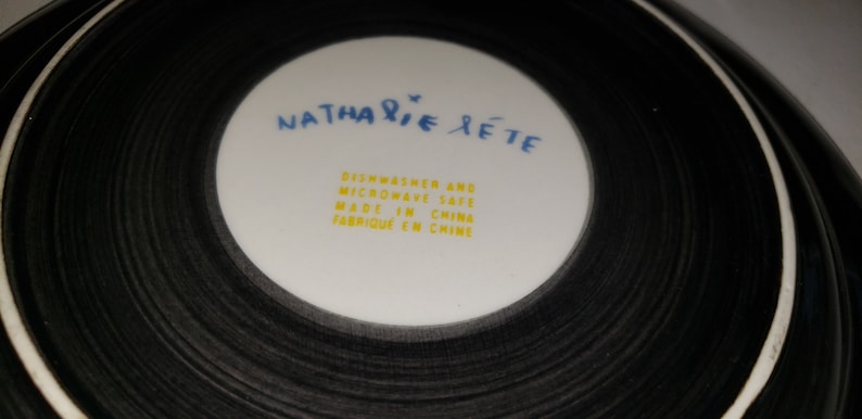May include: A white ceramic plate with black concentric circles around the edge. The center of the plate has the text "NATHALIE RETE" in blue and yellow text. The text "Dishwasher and Microwave Safe Made in China Fabrique en Chine" is printed in yellow.