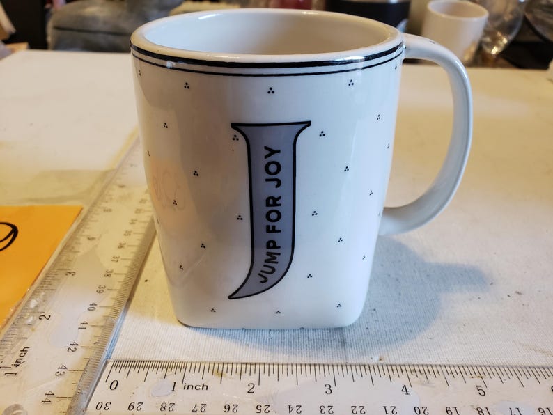K&ouml;nnte beinhalten: Wei&szlig;er Keramikbecher mit schwarzem Rand und Henkel. Der Becher hat einen gro&szlig;en grauen Buchstaben "J" mit den vertikal aufgedruckten Worten "JUMP FOR JOY". Kleine schwarze Punkte sind &uuml;ber die Oberfl&auml;che des Bechers verstreut. Ein Lineal ist im Vordergrund.