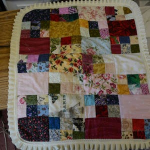 May include: A patchwork quilt with a white lace border. The quilt is made of various fabrics with floral and geometric patterns in shades of red, blue, green, and yellow.