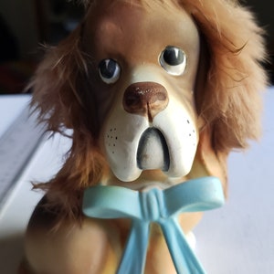 May include: A brown and white ceramic dog figurine with a blue bow. The dog has a furry brown head and a white face with black eyes and a black nose.