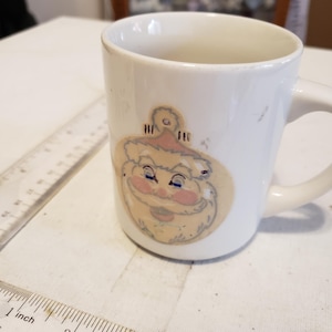 May include: White ceramic mug with a cartoon Santa Claus ornament design. The ornament is yellow, pink, and blue with a red hat.