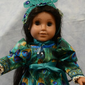 May include: A doll wearing a teal and blue peacock-patterned dress with a teal ribbon sash. The doll has long black hair and is wearing a teal and blue jeweled crown.