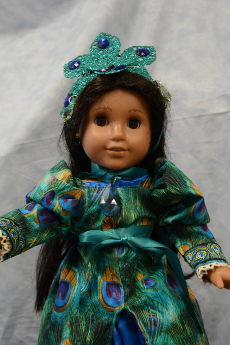 May include: A doll wearing a teal and blue peacock-patterned dress with a teal ribbon sash. The doll has long black hair and is wearing a teal and blue jeweled crown.