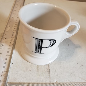 以下が含まれることがあります： 前面に黒い文字「P」が付いた白いセラミックマグ。マグにはハンドルと台座が付いています。マグの横には、センチメートルで測定する定規があります。