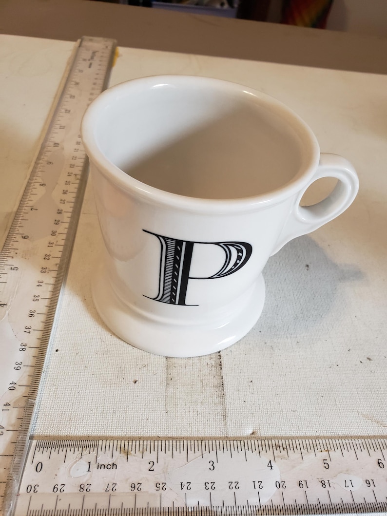 以下が含まれることがあります： 前面に黒い文字「P」が付いた白いセラミックマグ。マグにはハンドルと台座が付いています。マグの横には、センチメートルで測定する定規があります。