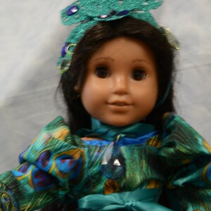 May include: A doll wearing a teal and green peacock-patterned dress with a teal sash and a jeweled headband. The doll has long black hair and brown eyes.