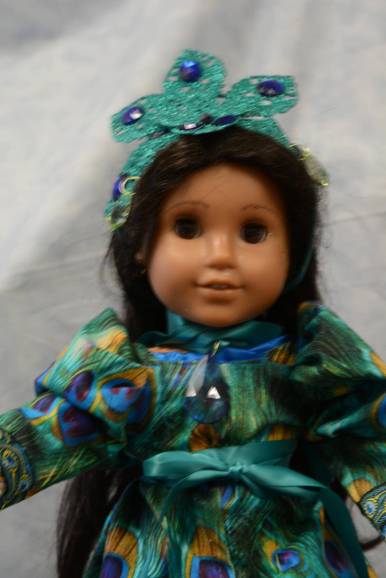 May include: A doll wearing a teal and green peacock-patterned dress with a teal sash and a jeweled headband. The doll has long black hair and brown eyes.
