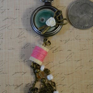 May include: A vintage-style necklace with a pocket watch pendant featuring a green and white design, a pair of scissors, and a white flower. The pendant is attached to a chain with a spool of pink thread and a tassel of beads and charms.