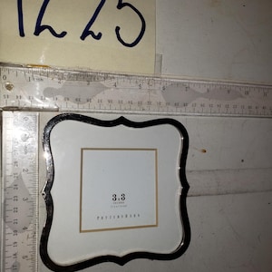 May include: A square Pottery Barn picture frame with a black border and a white interior. The frame is 3 x 3 inches (7.6 x 7.6 cm). The text "3x3 INCHES" and "POTTERY BARN" are printed on the frame.