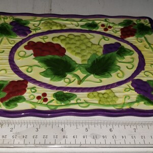 May include: A square ceramic trivet with a purple border and a design of green leaves, red and purple grapes, and a yellow background.