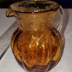 May include: A small amber glass pitcher with a textured surface and a clear glass handle.